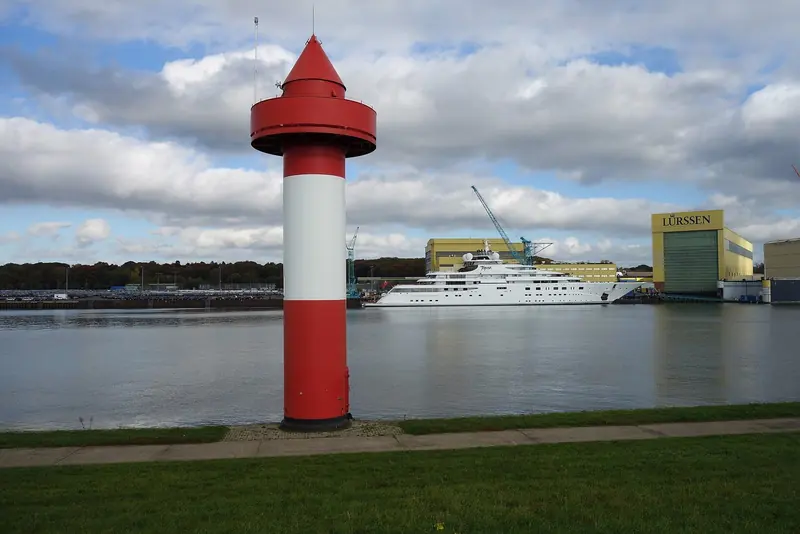 A large motor yacht lies at the Lurssen yard in Lemwerder, the shed doors and a harbour light marking the entrance to the fitting-out basin.