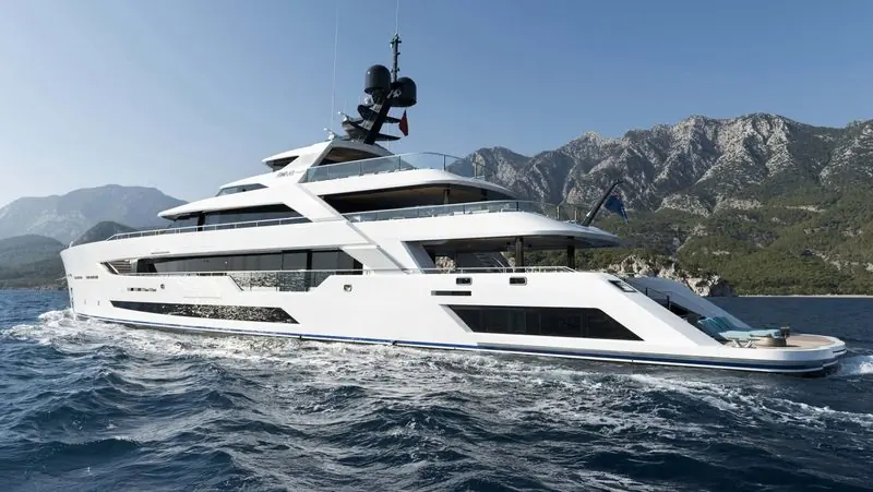 Mountains stand behind a white motor yacht as she runs along a rocky coast.