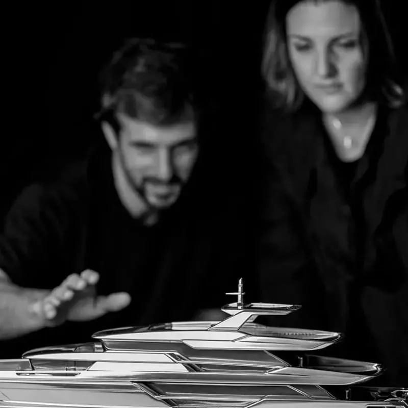 Two designers lean over a polished scale model of a motor yacht in a darkened studio.