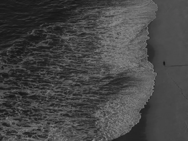 Waves fold onto an empty beach seen from directly above, a single walker's footprints trailing along the tideline.