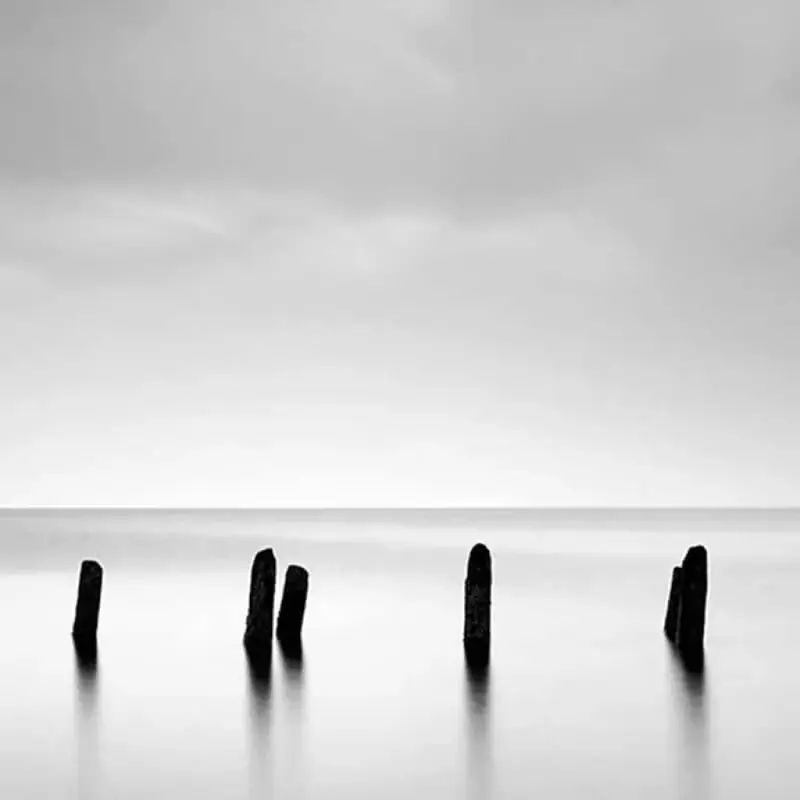Four weathered timber posts break a flat calm sea under a pale sky, all that remains of an old jetty.