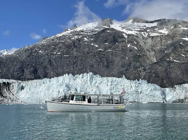 A small launch idles off the face of a tidewater glacier in Glacier Bay, giving the scale of the ice wall behind it.