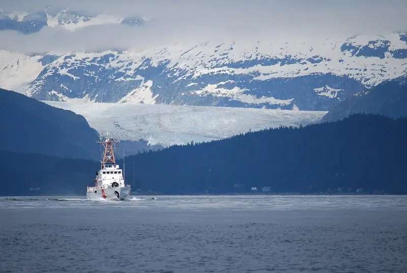 Under a glacier that fills the valley behind, a cutter runs down the channel near Juneau past houses along the shoreline.