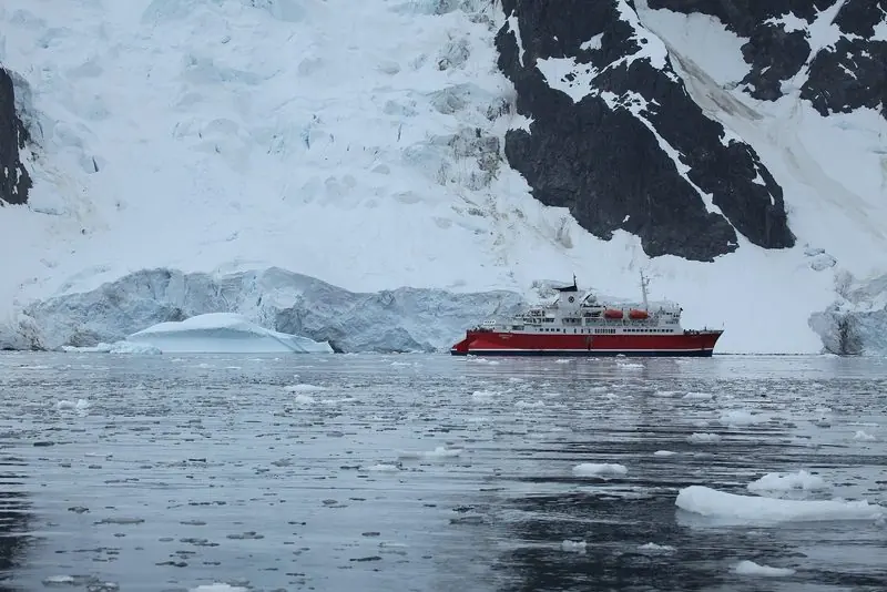An expedition ship lies close under a glacier front in loose brash ice, a grounded berg to one side and black rock above showing how steeply the shore falls away.
