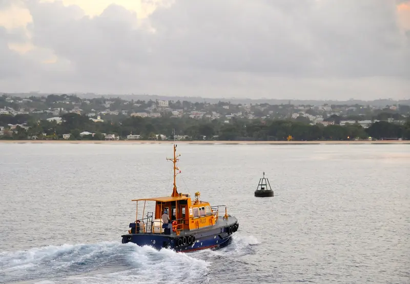 A harbour pilot boat runs out past the entrance buoy off Bridgetown, with the Barbados shoreline behind.