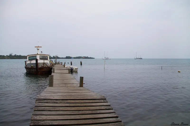 A working boat lies alongside a Placencia dock at day's end, two yachts anchored off in the lagoon beyond the last pilings.