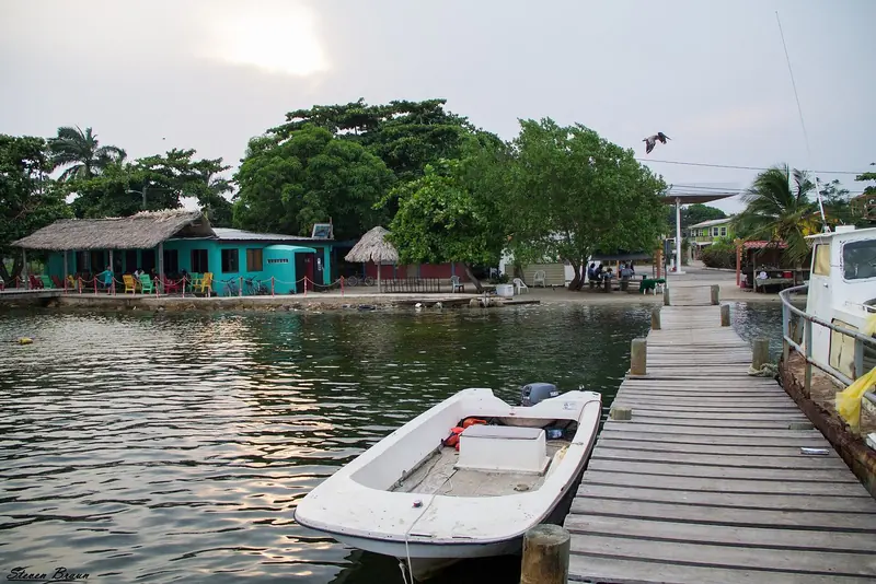 A skiff idles up to a Placencia dock past a thatched-roof boathouse, palms leaning into the wind off the water.