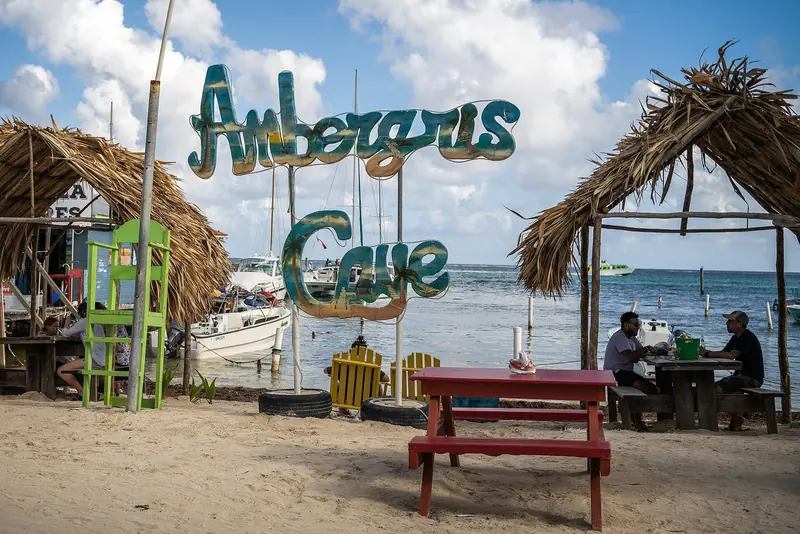 The Ambergris Caye sign stands over the San Pedro waterfront, boats moored on both sides of the beach bar where visitors gather.
