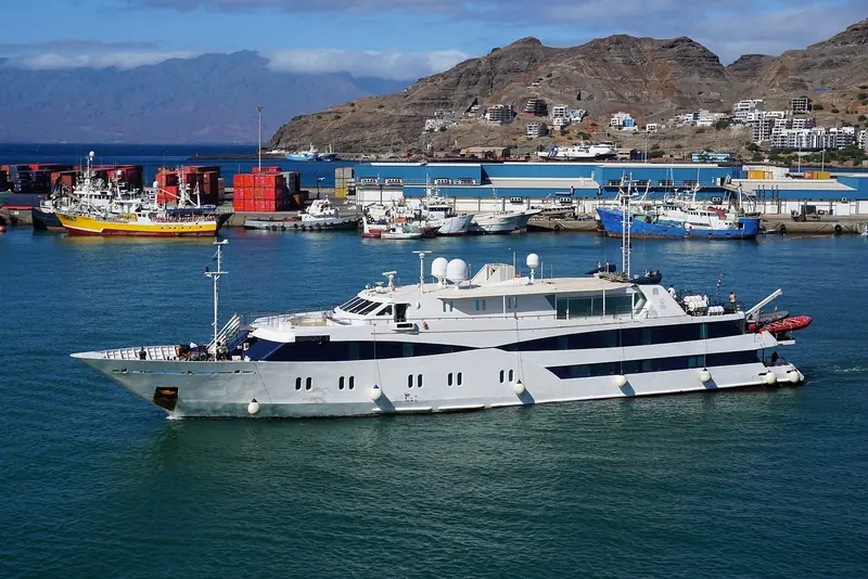 The 55m Harmony V departing Mindelo harbour past the town's fishing fleet, with Sao Vicente's volcanic ridgeline behind.