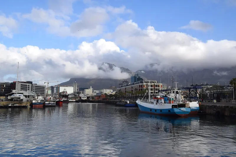 Fishing boats lie moored in the harbour with Table Mountain, half-covered in cloud, standing behind the city.