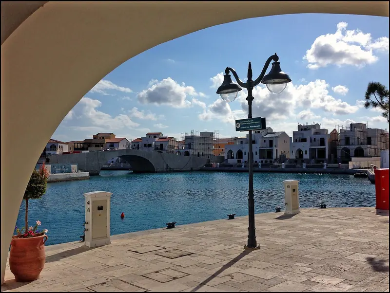 Limassol's waterfront promenade, with the marina and its apartment towers framed under an old stone arch.