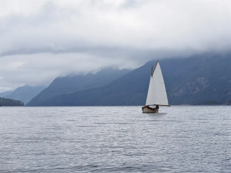 A gaff-rigged sailboat crosses Lewis Channel in the Discovery Islands, the cloud-wrapped mountains of the mainland shore behind it.