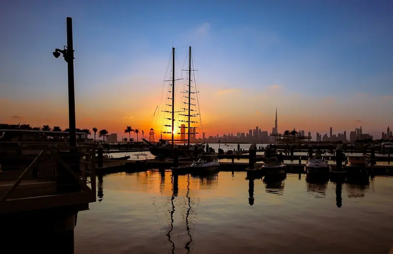 A large sailing yacht lies off at sunset with a marina in the foreground, in a season that runs October to May and closes in the summer heat.