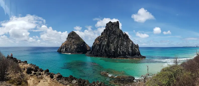 Morro Dois Irmaos (Two Brothers Peak) rising from the turquoise water at Baia dos Porcos, the volcanic landmark every arrival by sea or air uses to identify the island.
