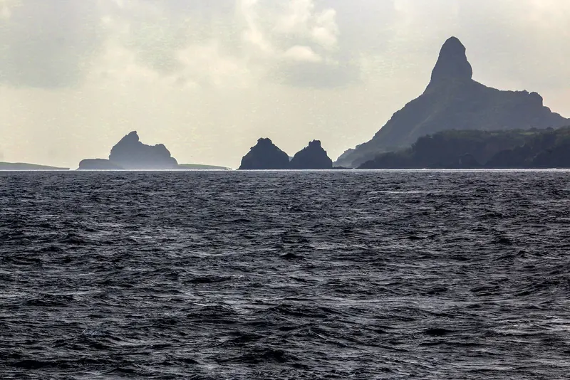 The archipelago's rock towers in silhouette as seen from a boat offshore, the same low, volcanic skyline that greets every vessel making the passage in from Recife or Natal.