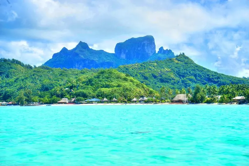 Bora Bora rises straight out of its lagoon, shallow turquoise water running unbroken across to a beach fringed with palms and thatched roofs.