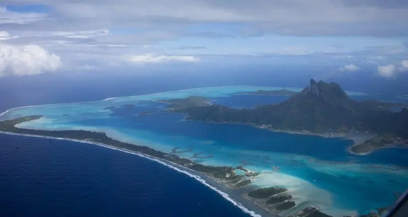 Seen from the air, the barrier reef encloses the whole island in graded blues, with motus, a pass and a line of surf marking where the ocean starts.