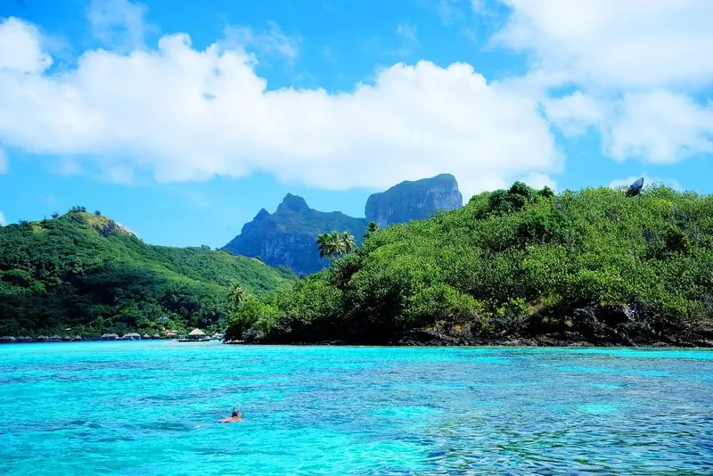 Clear shallow water runs up to a wooded motu with overwater bungalows off to one side and a swimmer working slowly along the coral.