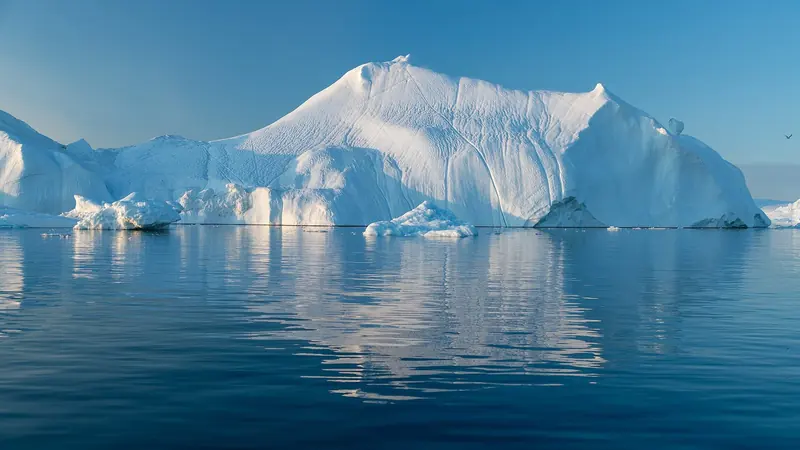 A sunlit iceberg calved from Sermeq Kujalleq drifts in the glass-calm water off Ilulissat, its underwater bulk visible in the reflection.