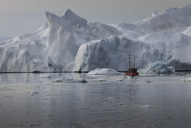 A local boat threads the water beneath a grounded iceberg near Ilulissat, the ice towering many times the vessel's height.