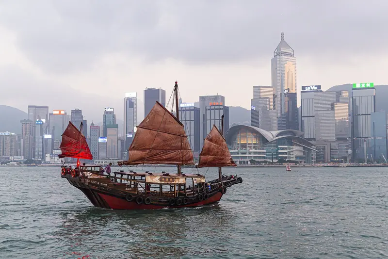 A red-sailed junk crosses Victoria Harbour at dusk against the Central skyline, the view most visitors associate with the harbour rather than with berthing in it.