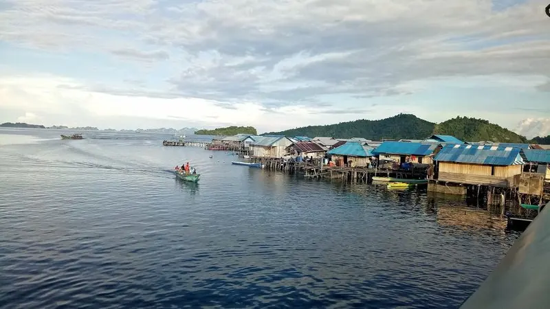 Houses on stilts run along the shore at a Misool village, with a longboat coming in and a larger craft standing off.