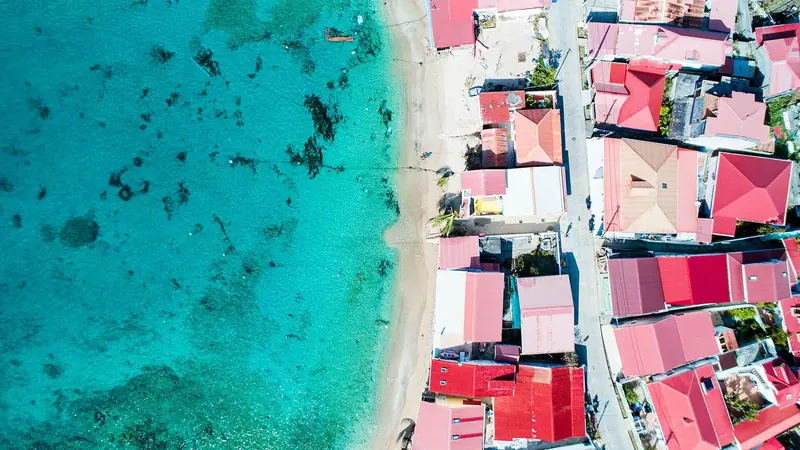 A vertical view over Les Saintes shows red-roofed houses lining a pale sand beach against clear turquoise shallows.