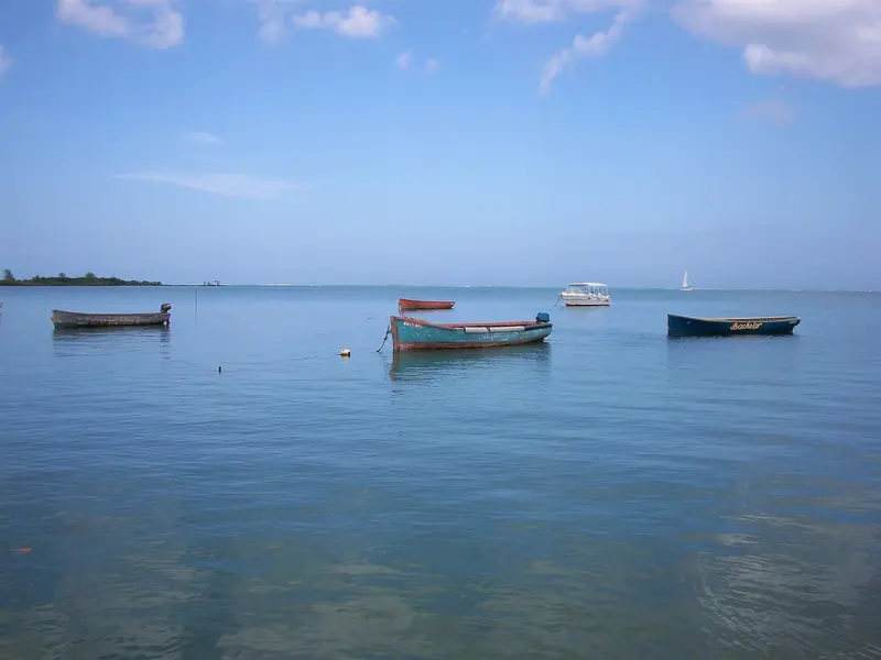 Small wooden pirogues and a day boat moored in a calm Mauritius lagoon, with a sailboat further out toward the reef line.