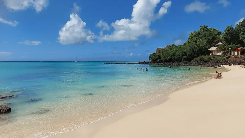 La Cuvette public beach at Grand Baie, swimmers in the clear lagoon with a yacht anchored offshore beyond the reef.