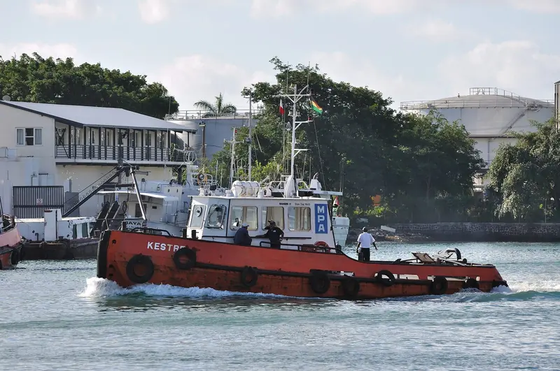 The Mauritius Ports Authority tug Kestrel underway in Port Louis harbour, crew on deck against the harbour's fuel tanks and warehouses.