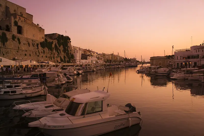 The harbour at Ciutadella at dusk, small craft rafted below the city walls with the narrow entrance channel running out to the west between them.