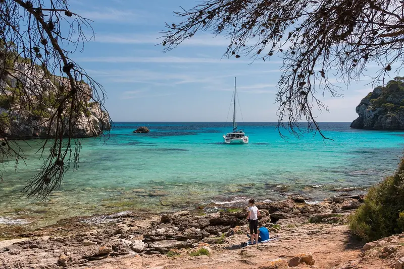 A sailing yacht and a catamaran on their hooks in Cala Macarella, framed by the pines that shade the cove's western headland.