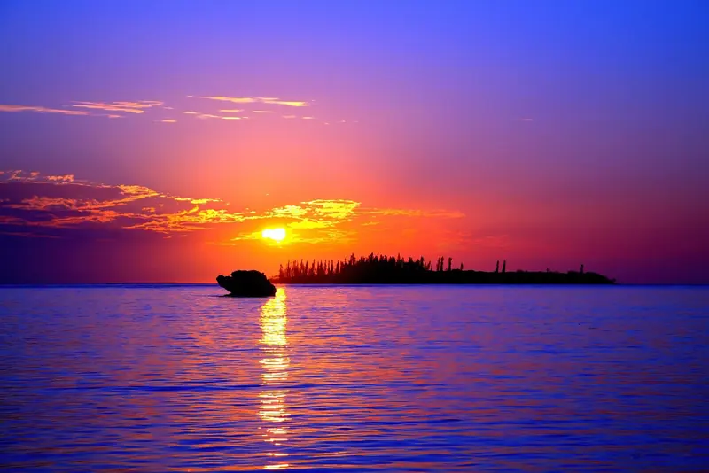 A small boat crosses the flat water off the Ile des Pins at sunset, the pines that gave the island its name silhouetted on the shore.