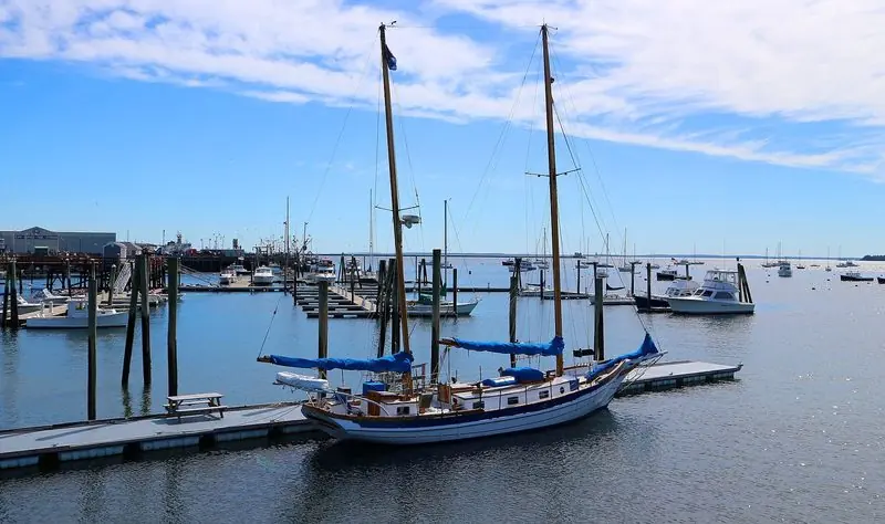 Two masts and blue sail covers mark a ketch alongside the float at Rockland, the fuel and service wharf close by and the open bay beyond.