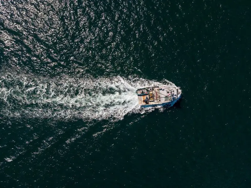 From directly above, a lobster boat lays a long white wake across dark green water, the working traffic that shares these bays.