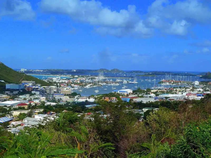 An aerial view over Simpson Bay Lagoon shows yacht and sailboat masts packed into the anchorage between Simpson Bay and Marigot, with the hills of St. Martin rising beyond the water.