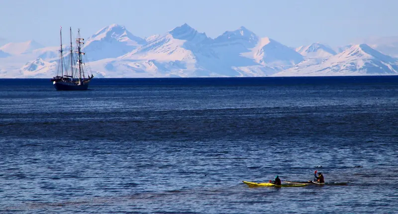 A sailing vessel lies off a snow covered range in Svalbard with a kayak alongside, in water that is open on the west coast from June.