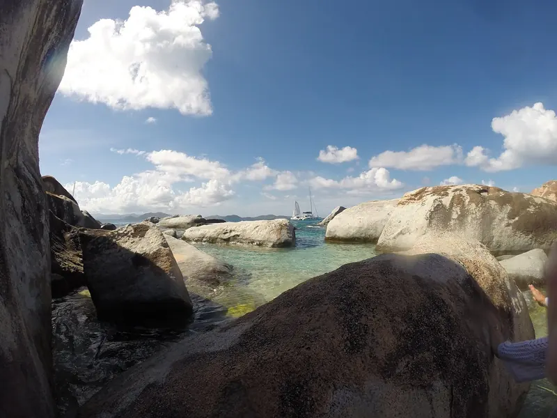 Granite boulders the size of houses at the Baths on Virgin Gorda, with a sailing yacht lying off the mooring field beyond them.