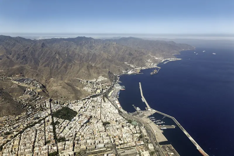 A Canarian coast from the air, with the port at the foot of a mountain wall and the trade wind running down the channel offshore.