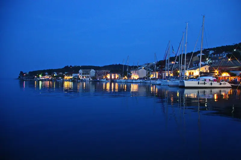 The harbour at Gaios on Paxos at dusk, with yachts stern to along the quay and the tavernas lit fifteen metres from the passerelle.