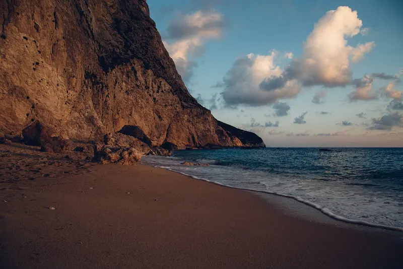 Late light on a Lefkada beach under the cliff, in a group that stays green through the summer because the Ionian takes real winter rain.