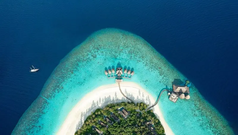 Seen from directly above, an island's reef runs from white sand through turquoise shallows into open blue, a two masted boat lying off the edge and villas built out along the flat.
