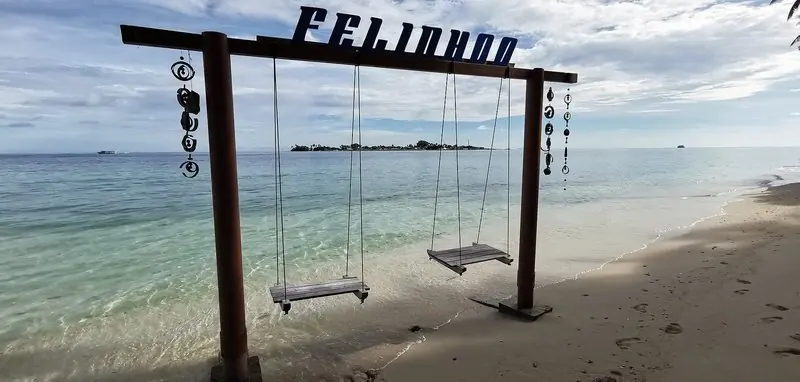 A swing frame planted at the tide line marks the landing beach on Felidhoo, with a neighbouring island low on the horizon across the channel.