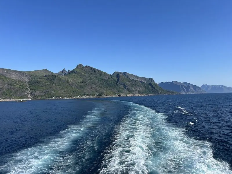 Wake curls away from the stern on the run between the Lofoten islands, a small settlement squeezed onto the shore below the mountains.