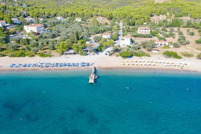 A yacht lies at anchor off Agioi Anargyri beach on Spetses, the water running from pale turquoise to deep blue over a sand bottom.