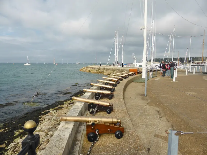 The line of brass cannon on the Cowes seafront, the most famous start line in sailing, with the masts of the yacht haven behind it.