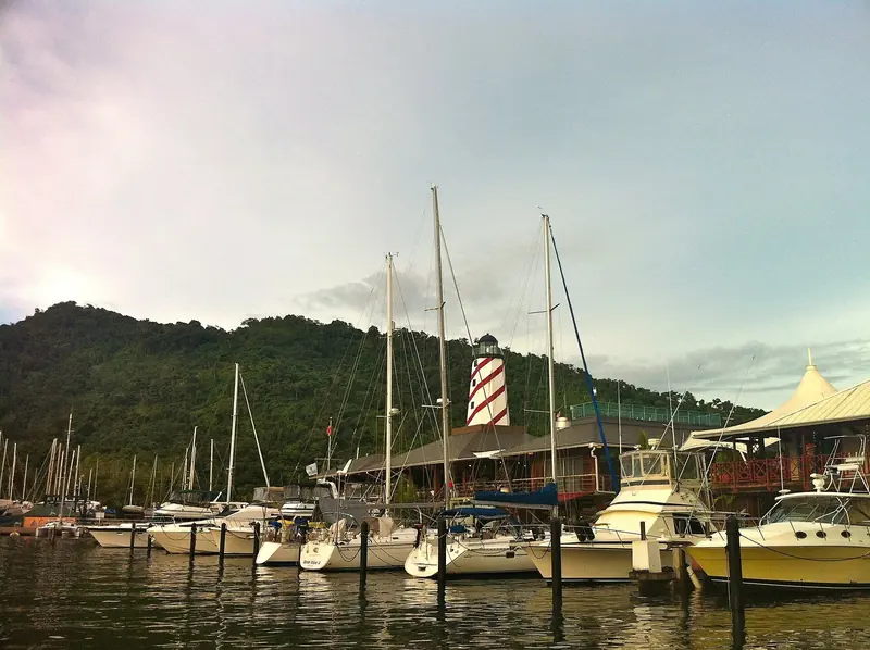 Crews Inn Marina at Chaguaramas, its red and white striped lighthouse marking the yachting hub on Trinidad's northwest peninsula, with cruising sailboats and sportfishers on the docks below the Northern Range.