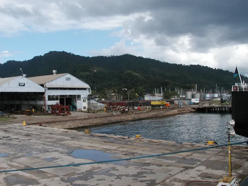A working boatyard on the Chaguaramas waterfront, plate shop and repair sheds backed by the forested hills of Trinidad's Northern Range, with yacht masts visible at the yards further along the bay.