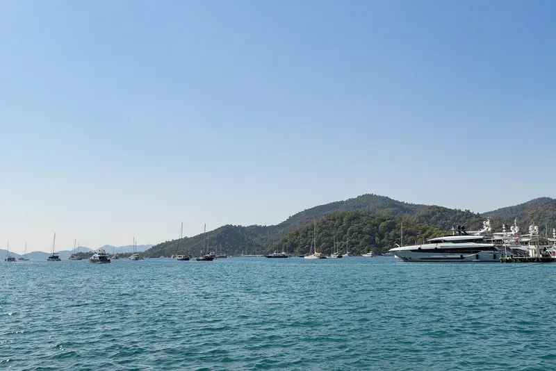Gocek Bay opens wide and deep behind low pine ridges, a motoryacht lying alongside the quay on the right and a scattered fleet swinging in flat water.