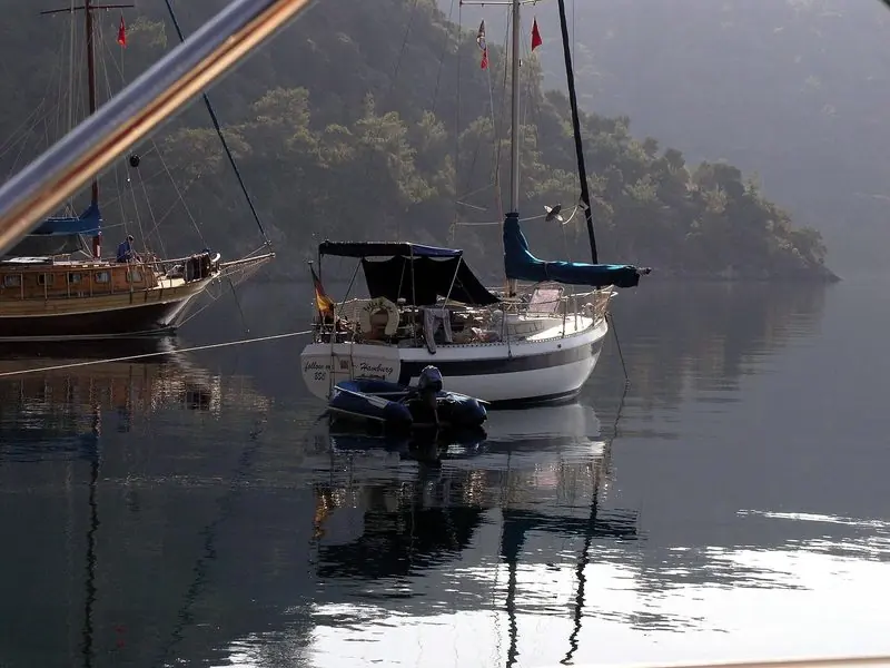 Early haze holds the anchorage still, a gulet and a small sloop lying to lines run ashore under a steep wooded slope with tenders tied astern.
